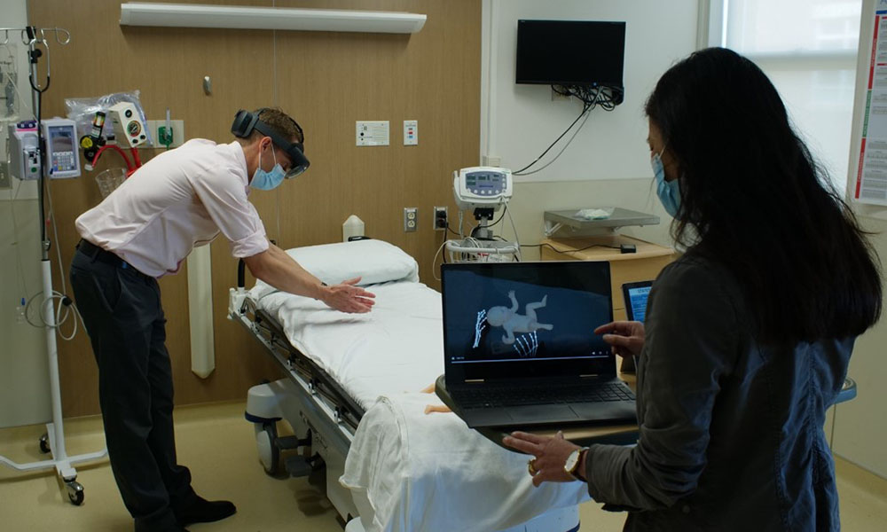 a man leaning over a hospital bed demonstrating the augmented reality simulation of a newborn in distress.