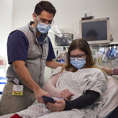 a doctor wearing a mask standing next to a patient on a gurney in an emergency room setting. Both are looking at the patient's cellphone.