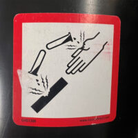 Close-up view of a hazard sticker indicating a chemical that can burn or injure surfaces and skin